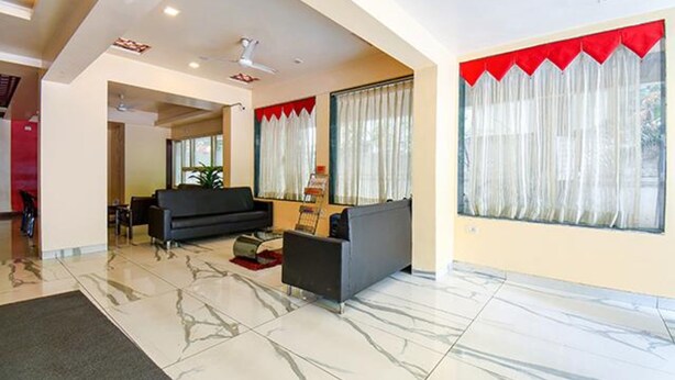 A spacious living room with a black sofa, a coffee table, and a TV. There are large windows with red curtains and a view of the outdoors. - Fabhotel Envirosafe Stays (Pune)