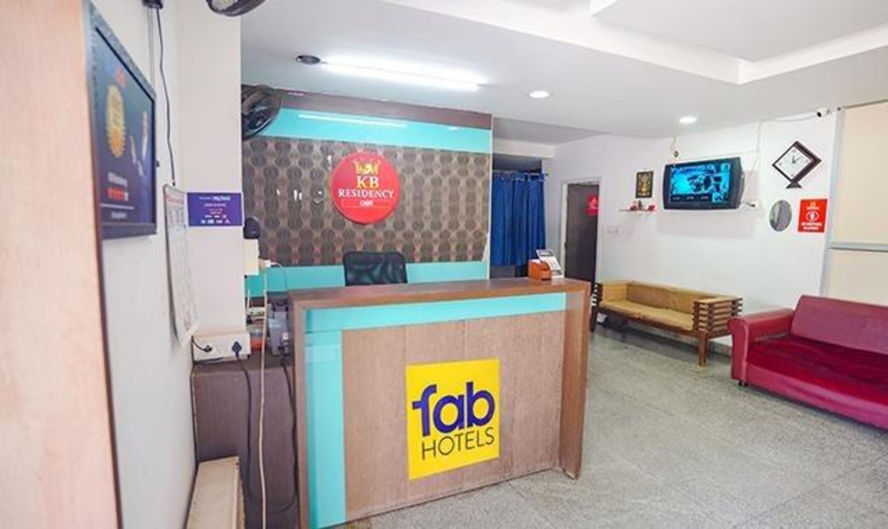 Zona de recepción de un hotel con un mostrador de recepción que dice 'fab hotels'. Hay un sofá rojo y un televisor montado en la pared.