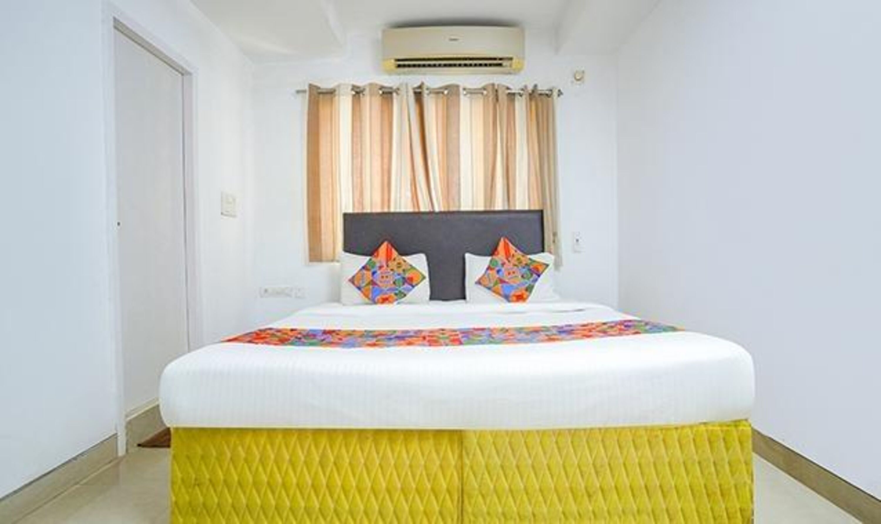 A hotel room with a bed, curtains, an air conditioning unit, and a window.