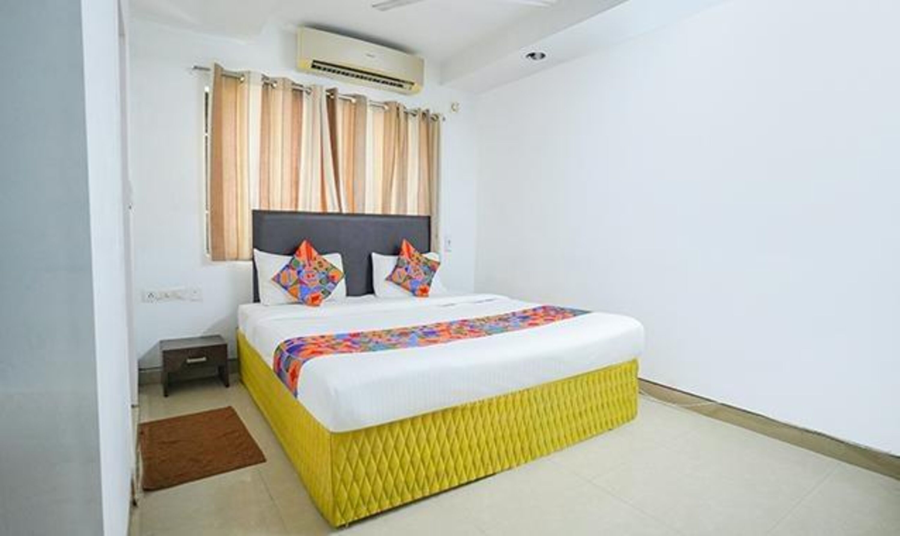 A hotel room with a bed, a nightstand, an air conditioning unit, and a window with curtains.
