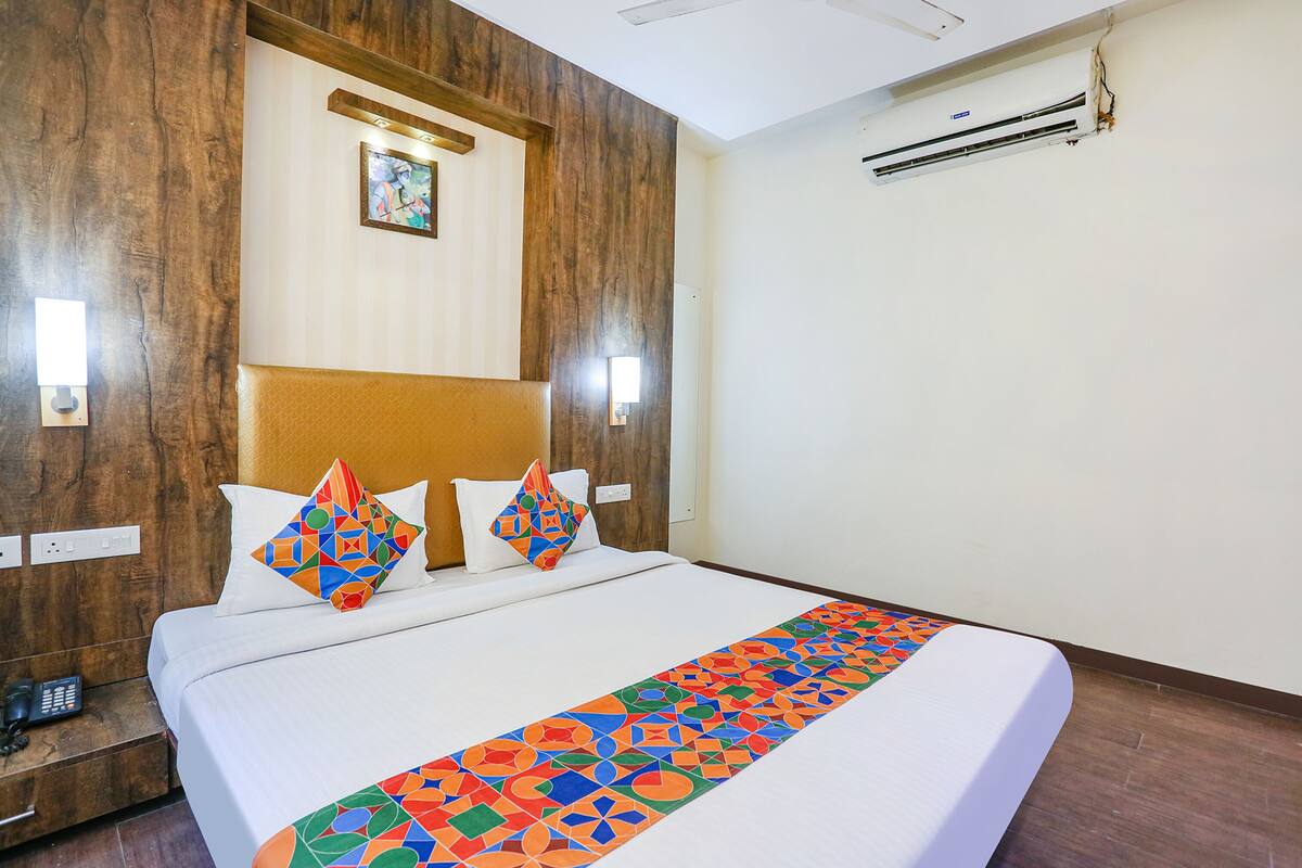 A hotel room with a large bed, two pillows with geometric patterns, a wooden headboard, a wall-mounted air conditioner, and a small painting above the headboard.