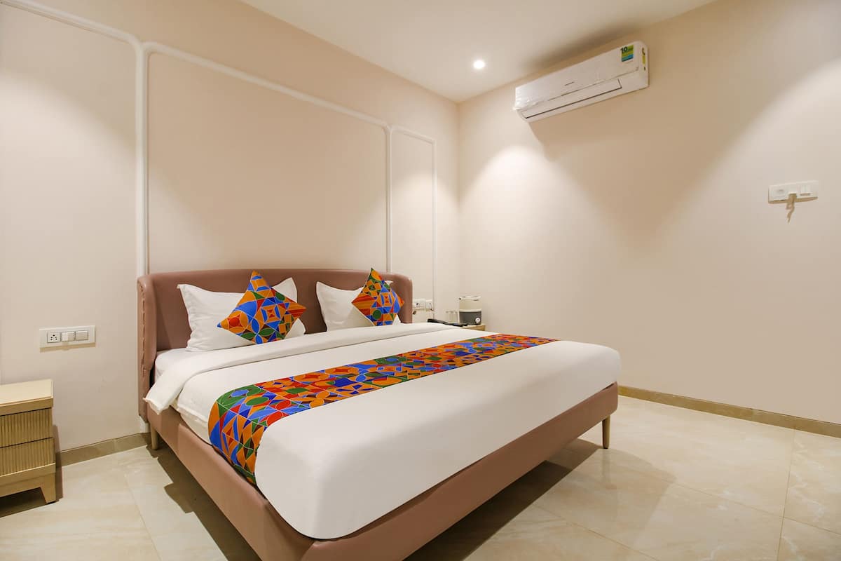 Un lit bien fait, recouvert d’une couvre-lit aux motifs géométriques colorés, une table de chevet avec une lampe et un appareil de climatisation fixé au mur.
