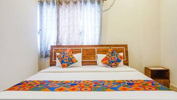 A hotel room with a bed, wooden headboard, colorful pillows, a patterned bedspread, and a window with curtains.