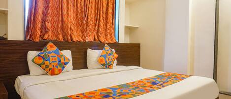 A hotel room with a neatly made bed, a wooden headboard, and a window with orange curtains.