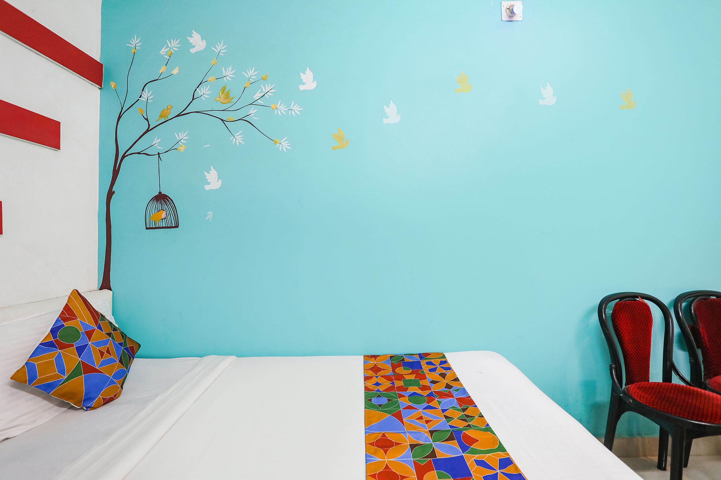 Ein Zimmer mit einem Bett, einem farbenfrohen Kissen, einer Bettdecke mit geometrischem Muster, einer Wandmalerei mit Baum und Vögeln sowie zwei roten Stühlen.