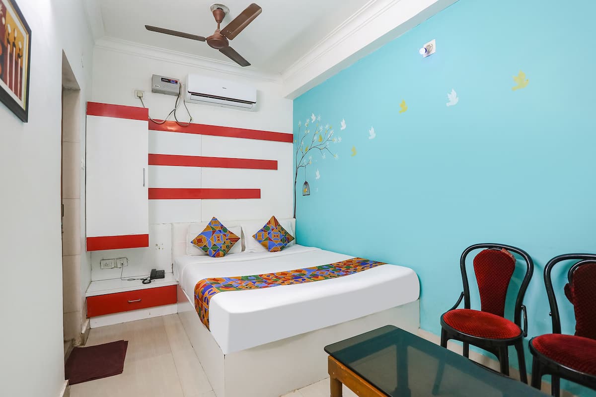 Une chambre avec un lit, un ventilateur de plafond, un appareil de climatisation, un mur décoré d’une fresque représentant un arbre, et deux chaises rouges.