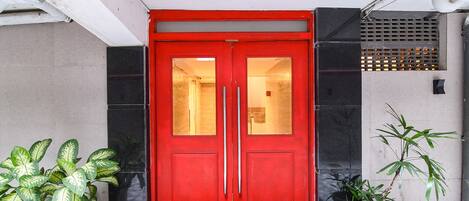 Une porte rouge à deux battants avec des panneaux de verre, un pot de fleurs sur le côté droit et un mur recouvert de carreaux.