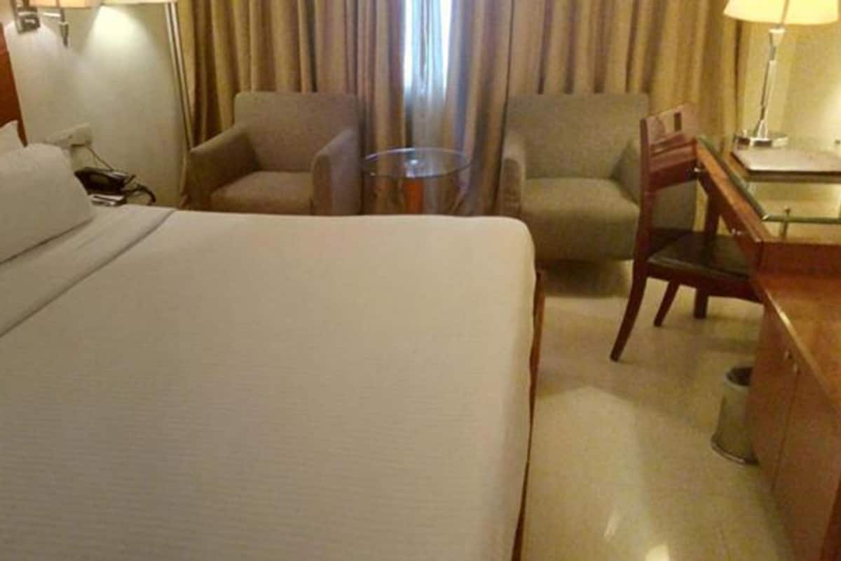 Quarto de hotel com uma cama grande, duas poltronas, uma mesinha e uma escrivaninha com abajur.