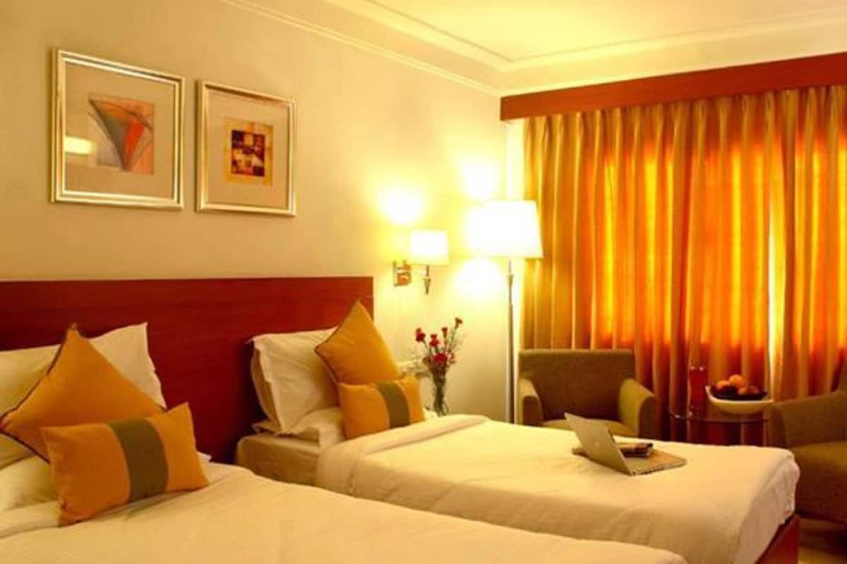 Quarto de hotel com duas camas, uma escrivaninha, uma cadeira e uma luminária.