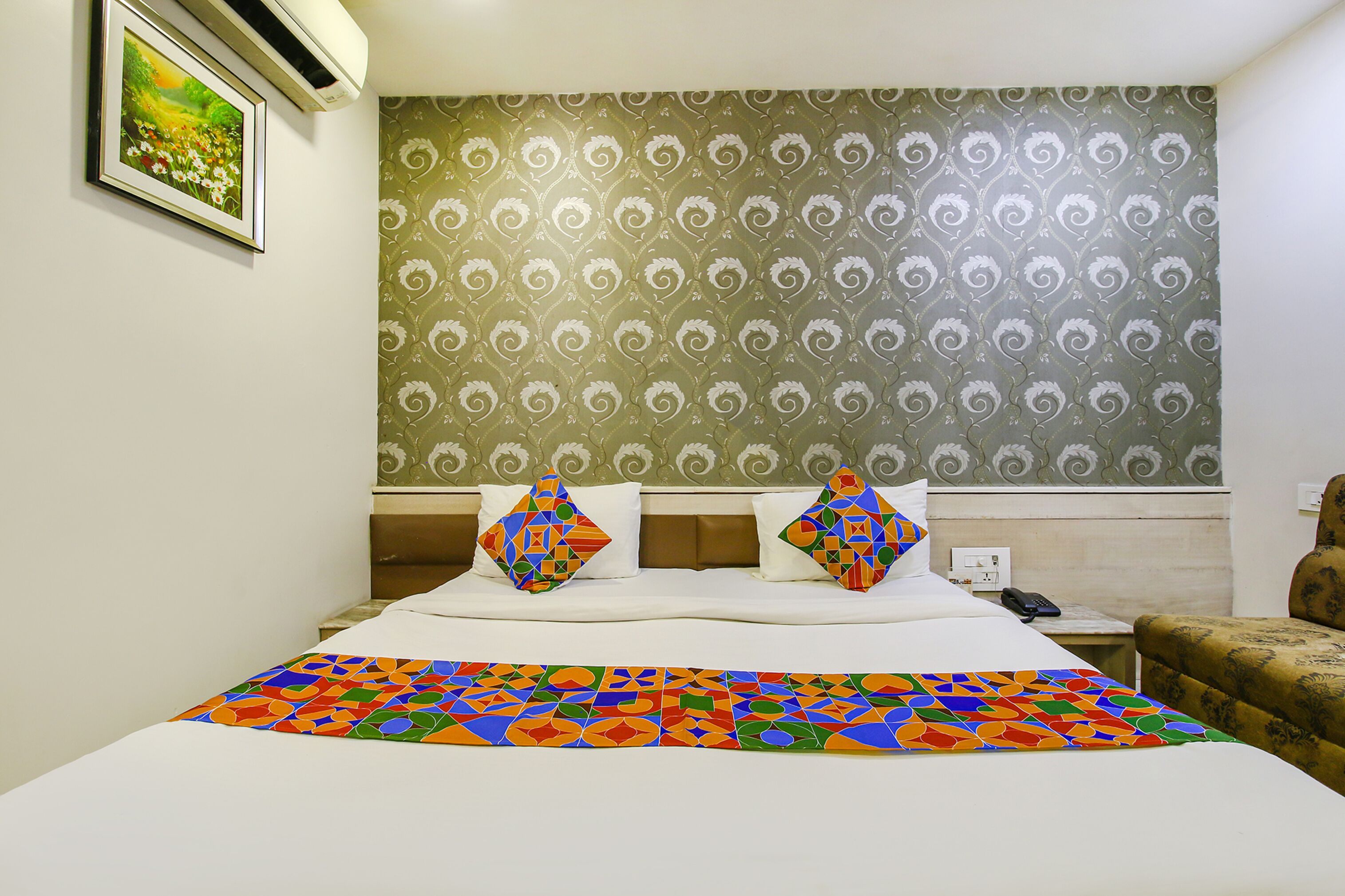 A hotel room with a bed, two pillows, a patterned wallpaper, a framed picture on the wall, and a nightstand with a phone.