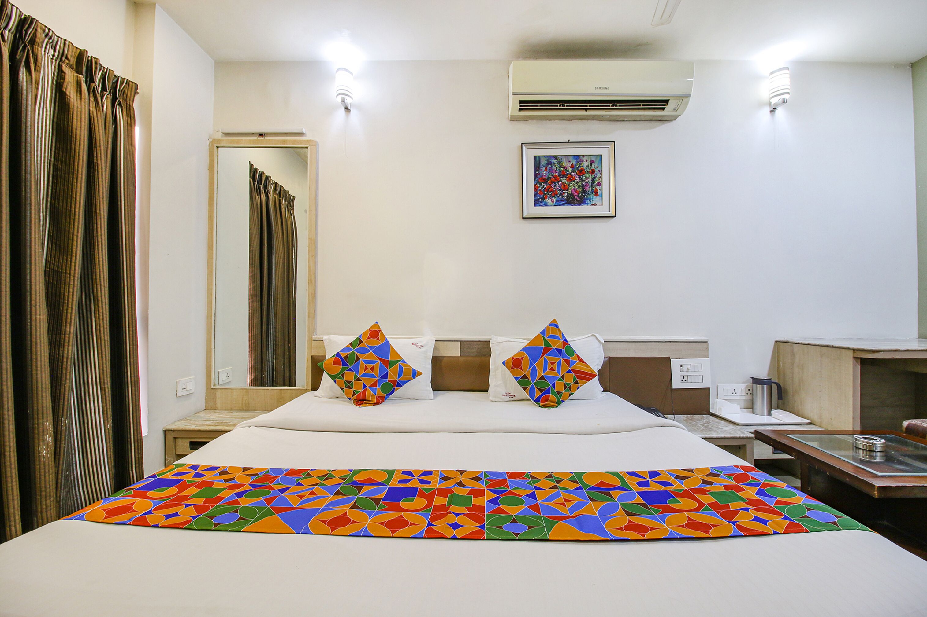 A hotel room with a bed, a bedside table, a mirror, an air conditioner, and a painting on the wall.