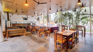 A restaurant with wooden furniture, ceiling fans, and large windows with a view of palm trees. - Oriva Shourya Residency (Pune)