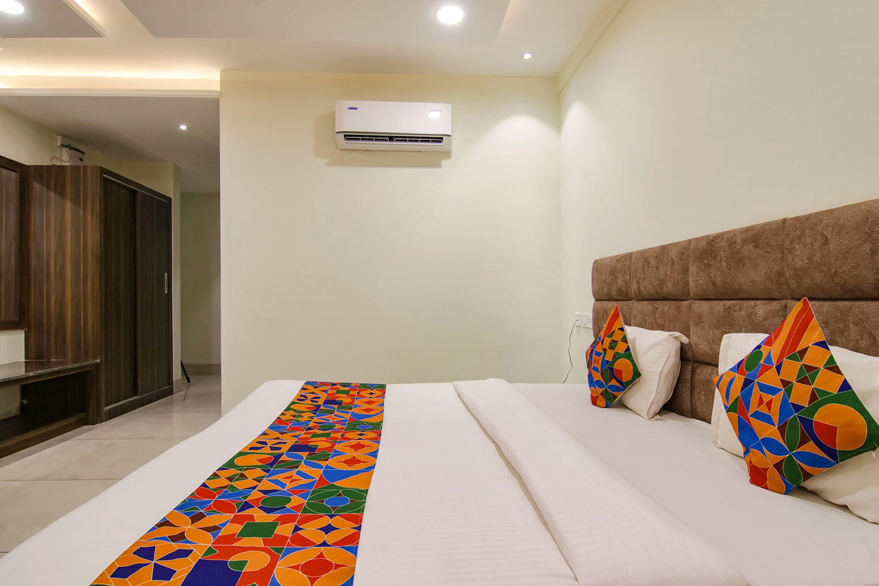 Quarto de hotel com cama, um tapete de cor vibrante com padrão geométrico, cabeceira marrom e unidade de ar condicionado.
