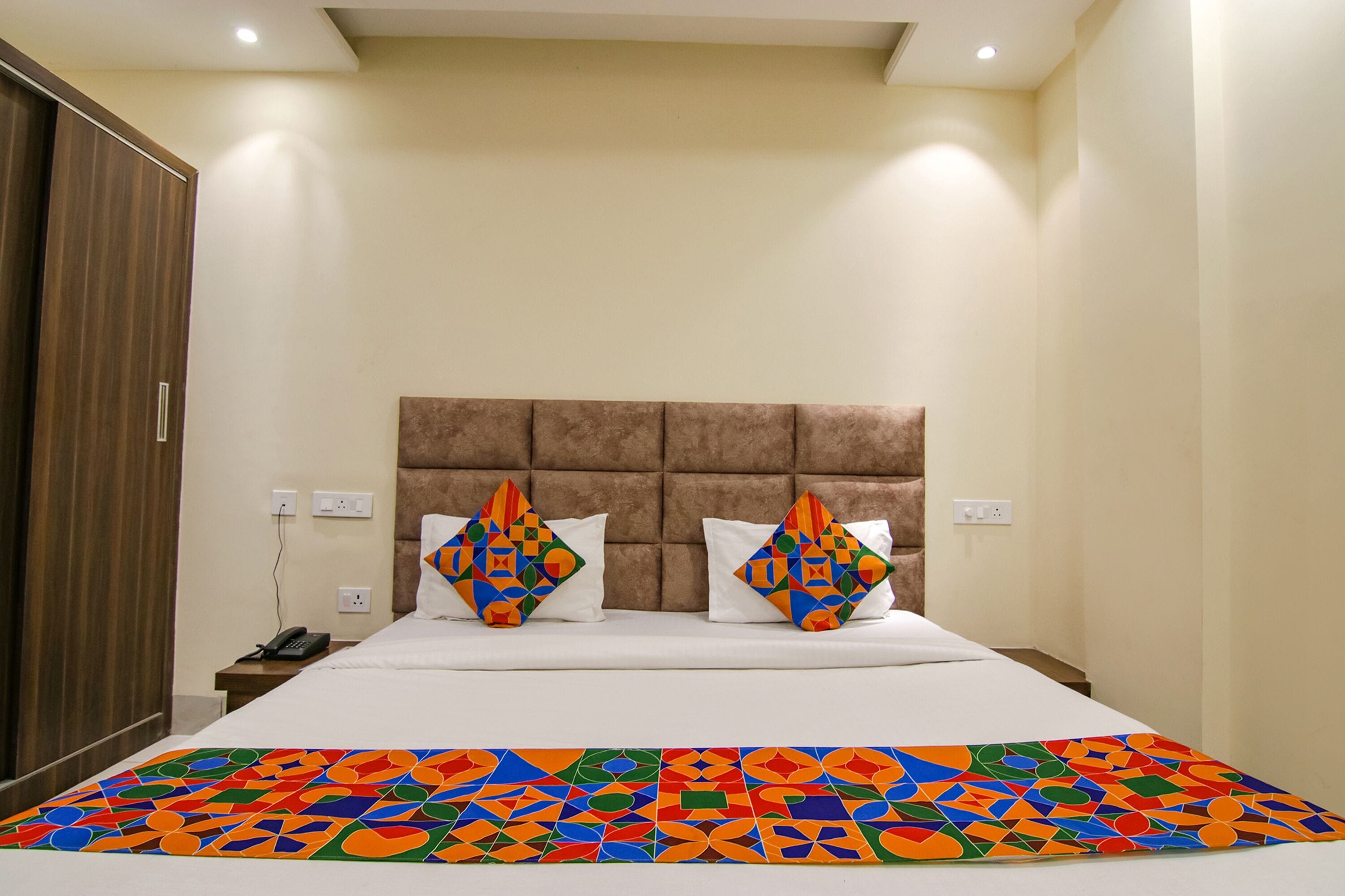 A hotel room with a large bed, a headboard, two pillows with a geometric pattern, a bedside table with a phone, and a colorful bedspread.