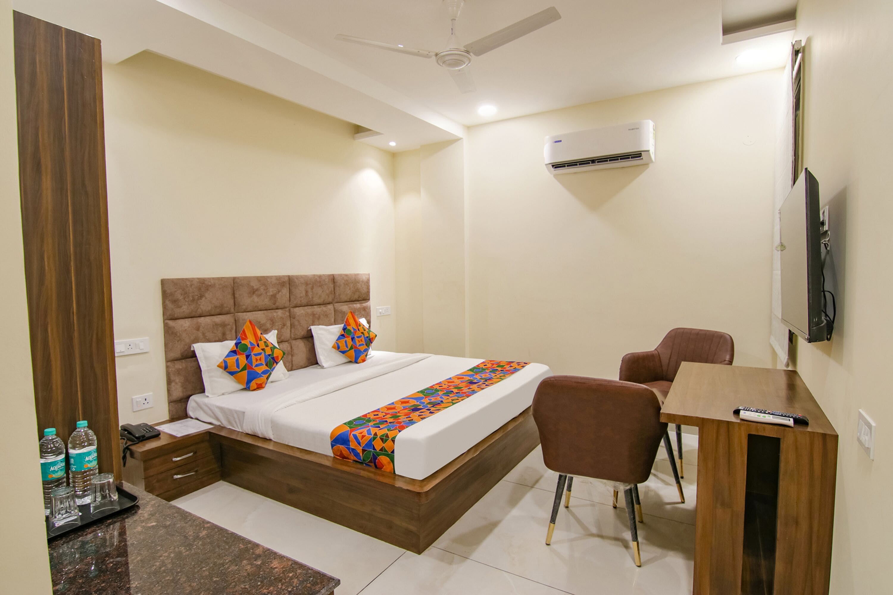 A hotel room with a bed, a desk, a chair, a TV, and an air conditioning unit.