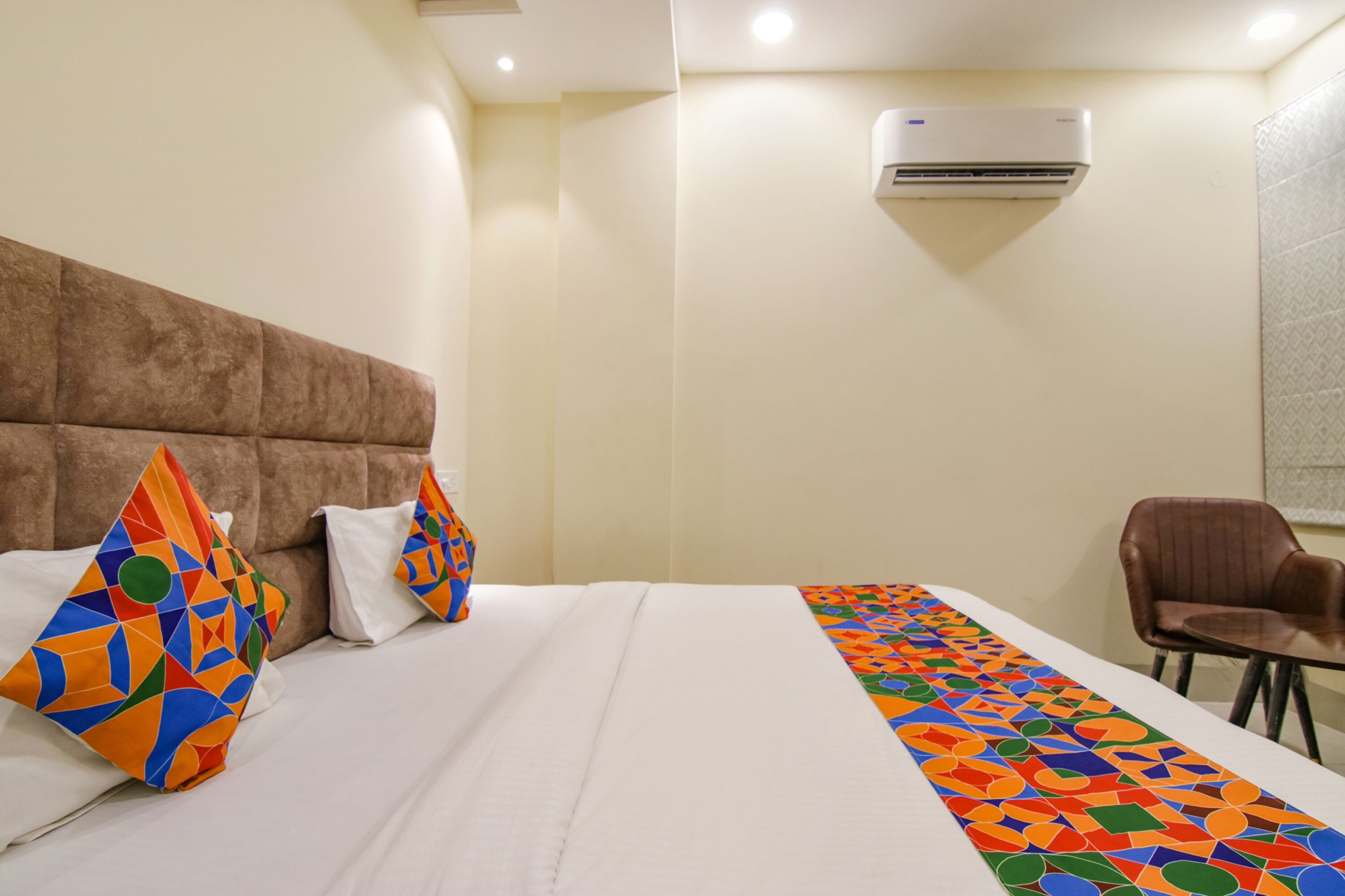 Habitación de hotel con una cama grande, cabecera marrón, colcha estampada, una silla marrón y un equipo de aire acondicionado en la pared.