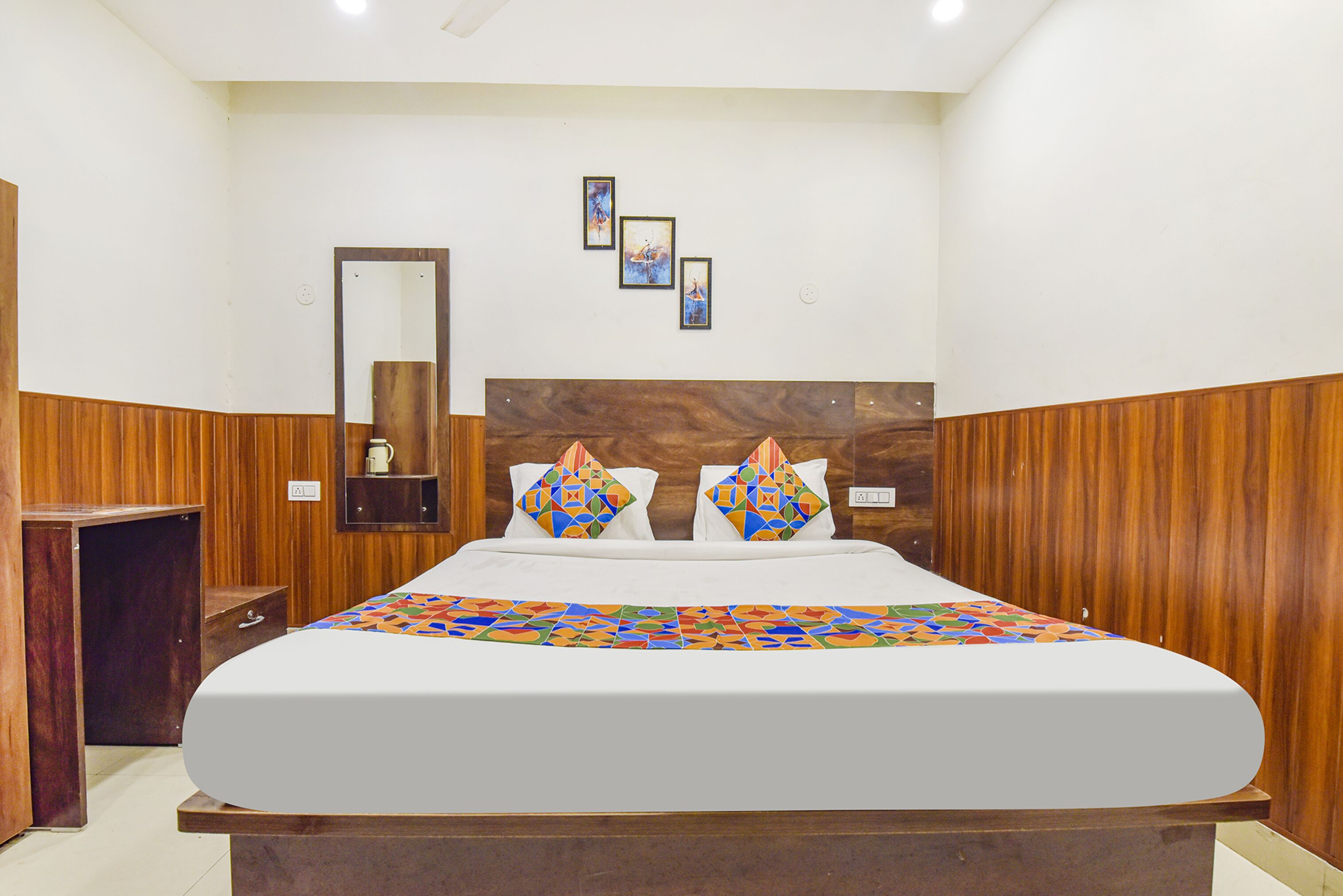 Un dormitorio ordenado con cabecera de madera, cama con colcha de estampado geométrico y colorido, dos almohadas decorativas, una mesita de noche y un espejo montado en la pared.