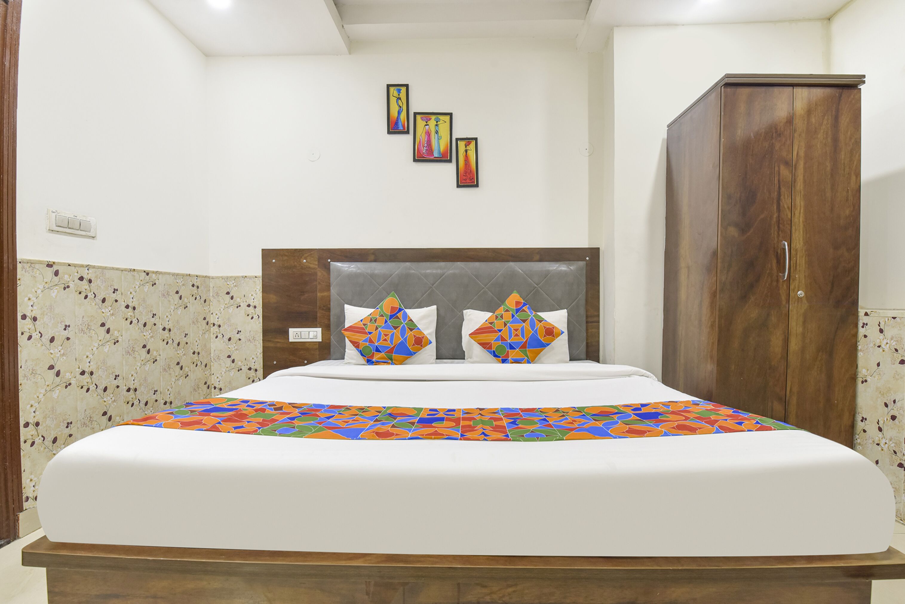 Un lit bien fait, agrémenté d’une couvre-lit aux motifs géométriques colorés, d’une tête de lit en bois et d’une armoire en bois, dans une pièce aux murs blancs et dont les murs sont décorés d’une bordure de papier peint à motifs floraux.