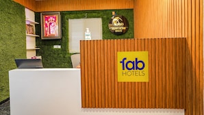 A reception area with a wooden reception desk, a green wall with artificial grass, a framed picture, a window with blinds, and a sign that reads "fab hotels". - FabHotel Happy Home Stay (Bengaluru)