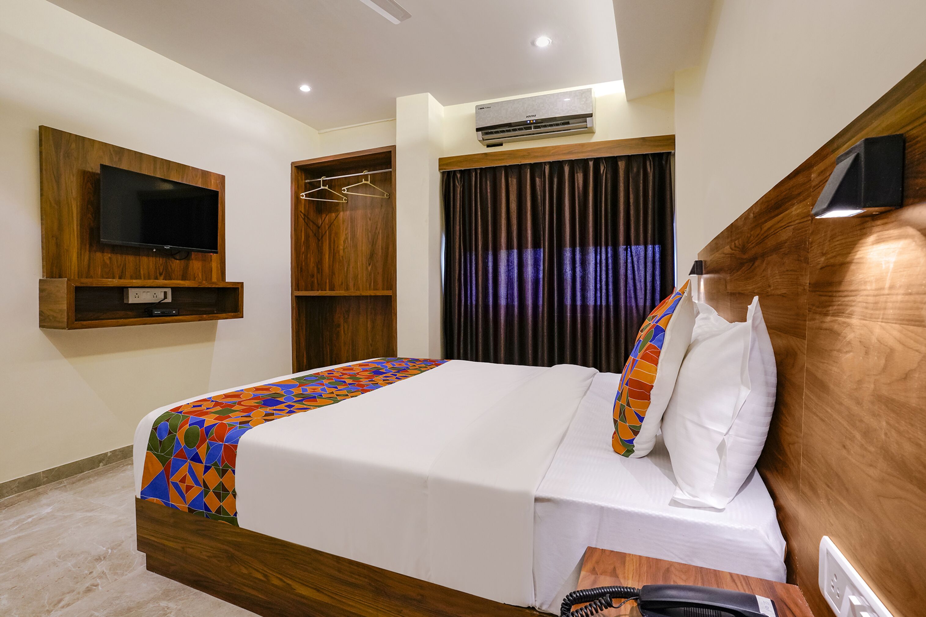 A hotel room with a bed, a TV mounted on the wall, a wardrobe, and an air conditioning unit.