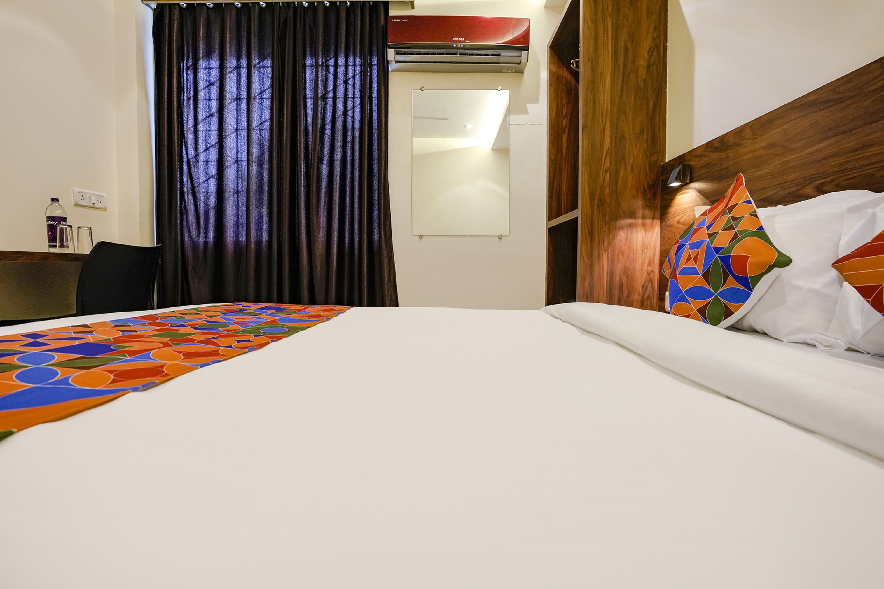 A hotel room with a bed, a desk, a chair, a window with curtains, and an air conditioning unit.