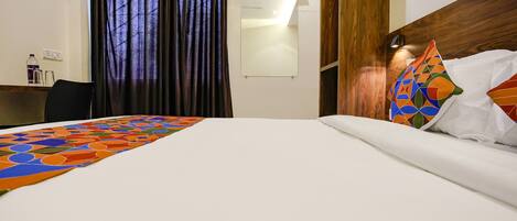 A hotel room with a bed, a desk, a chair, a window with curtains, and an air conditioning unit.