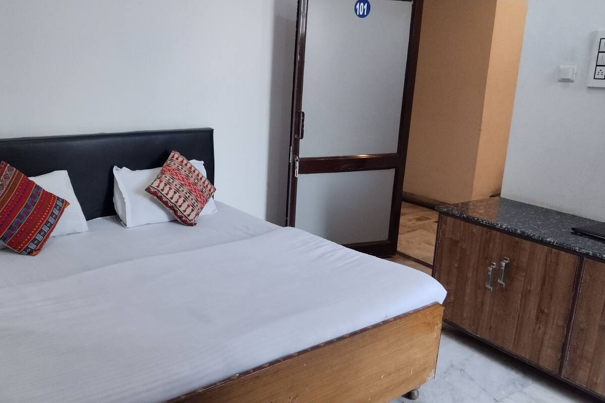 A neatly made bed with white linens and two decorative pillows, a wooden headboard, a wooden nightstand, and a wooden wardrobe with a marble countertop.