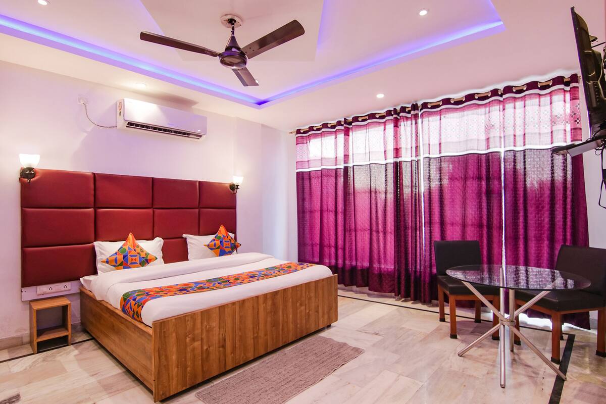 A modern bedroom with a wooden bed, a ceiling fan, an air conditioner, a glass table with chairs, and a window with purple curtains.