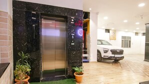A hotel entrance with a marble wall, an elevator, a white car parked outside, and potted plants. - Fabhotel Grover (Kangra)