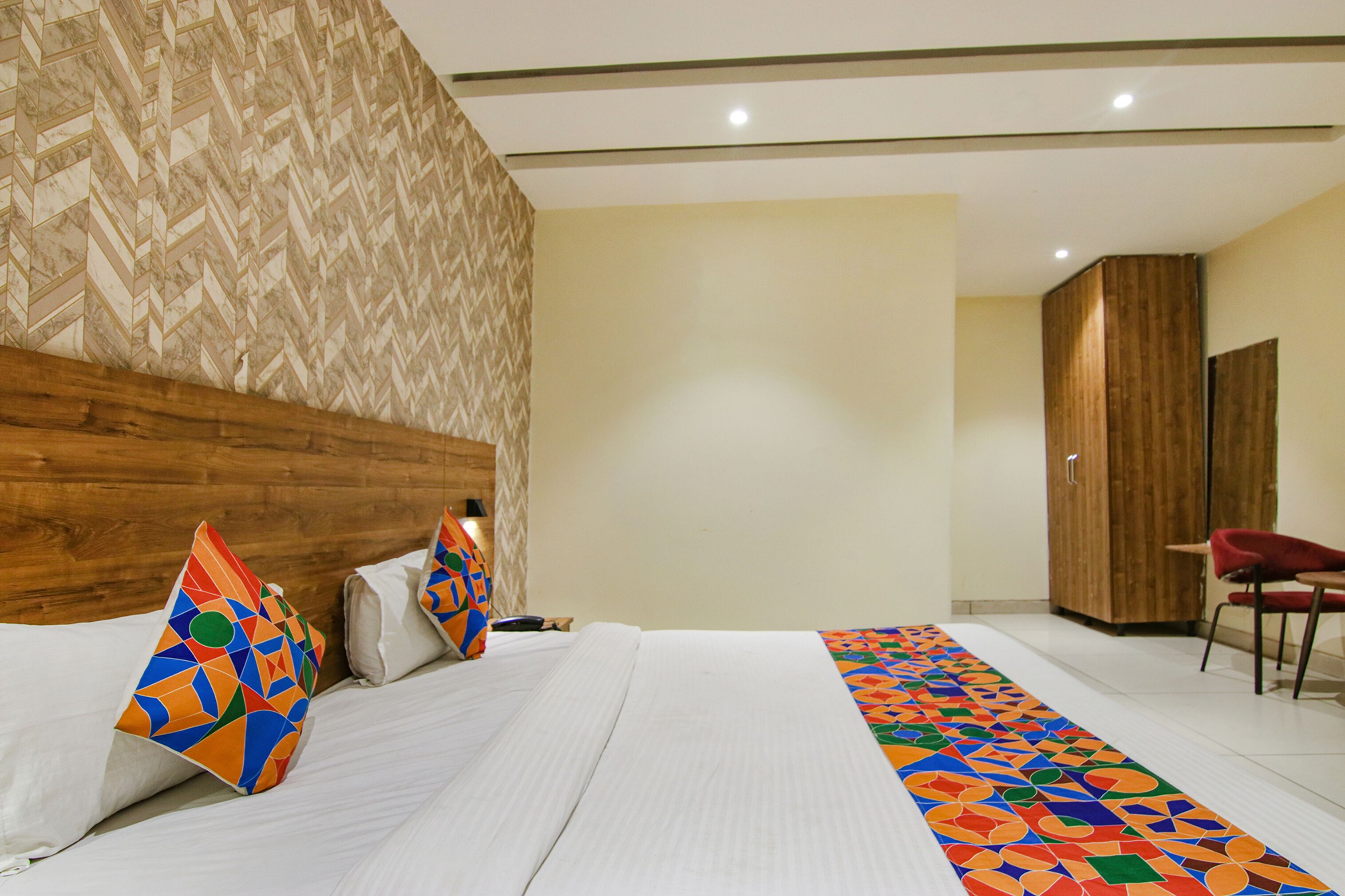 A hotel room with a wooden headboard, a large bed with patterned pillows, a colorful runner, and a wardrobe.