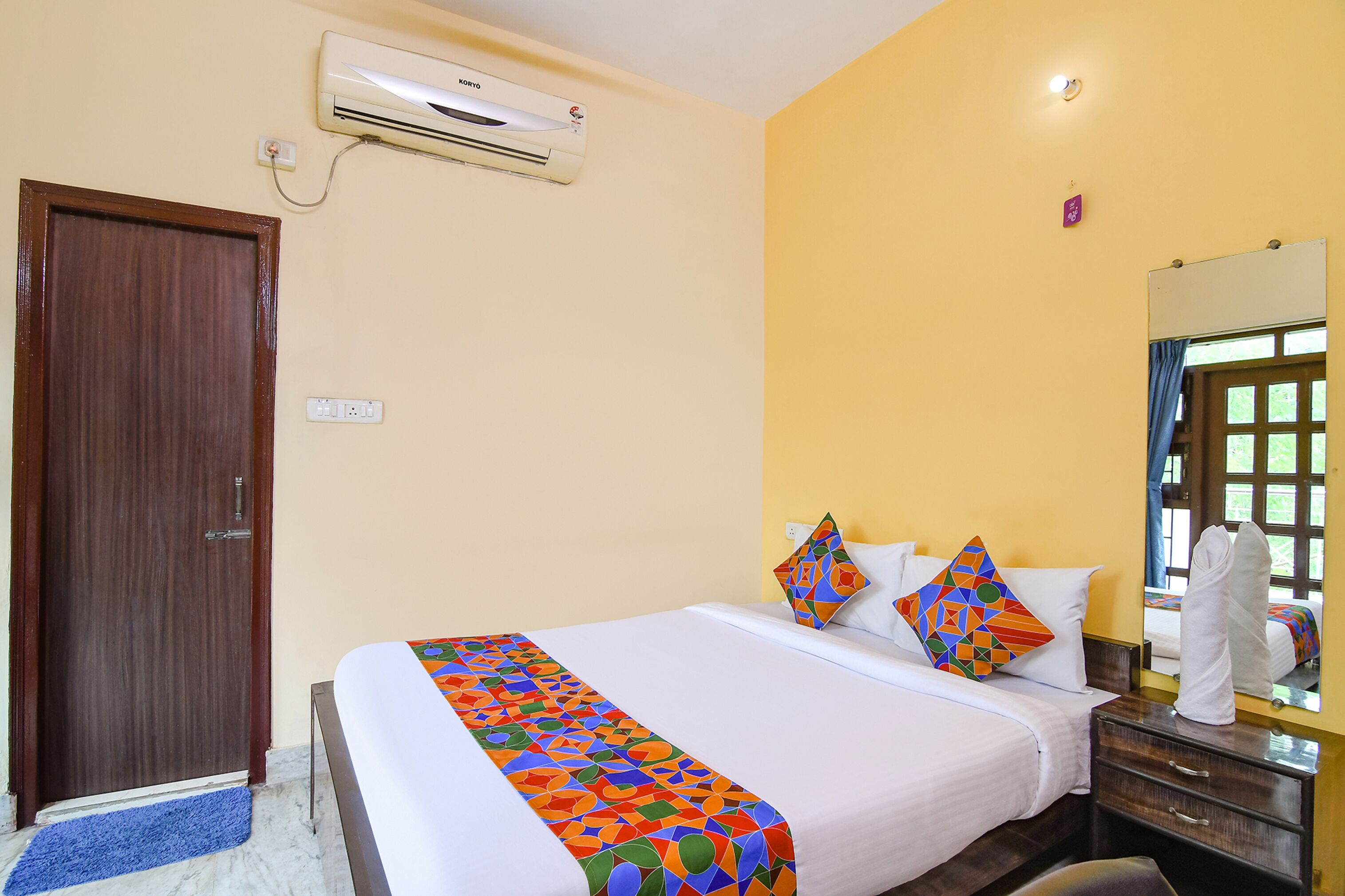 A hotel room with a bed, a wooden door, an air conditioning unit, a bedside table, and a mirror.