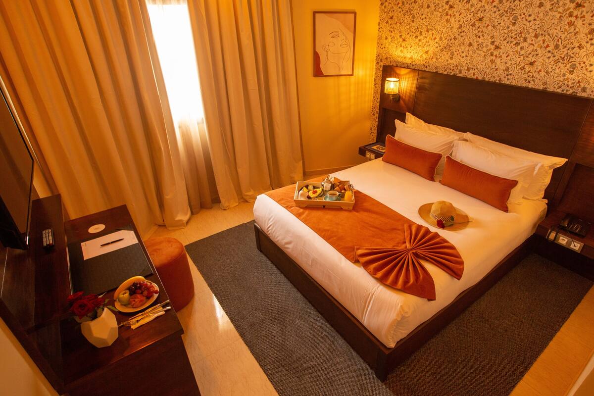 A hotel room with a bed, a bedside table, a lamp, a tray with breakfast items, and a window with curtains.