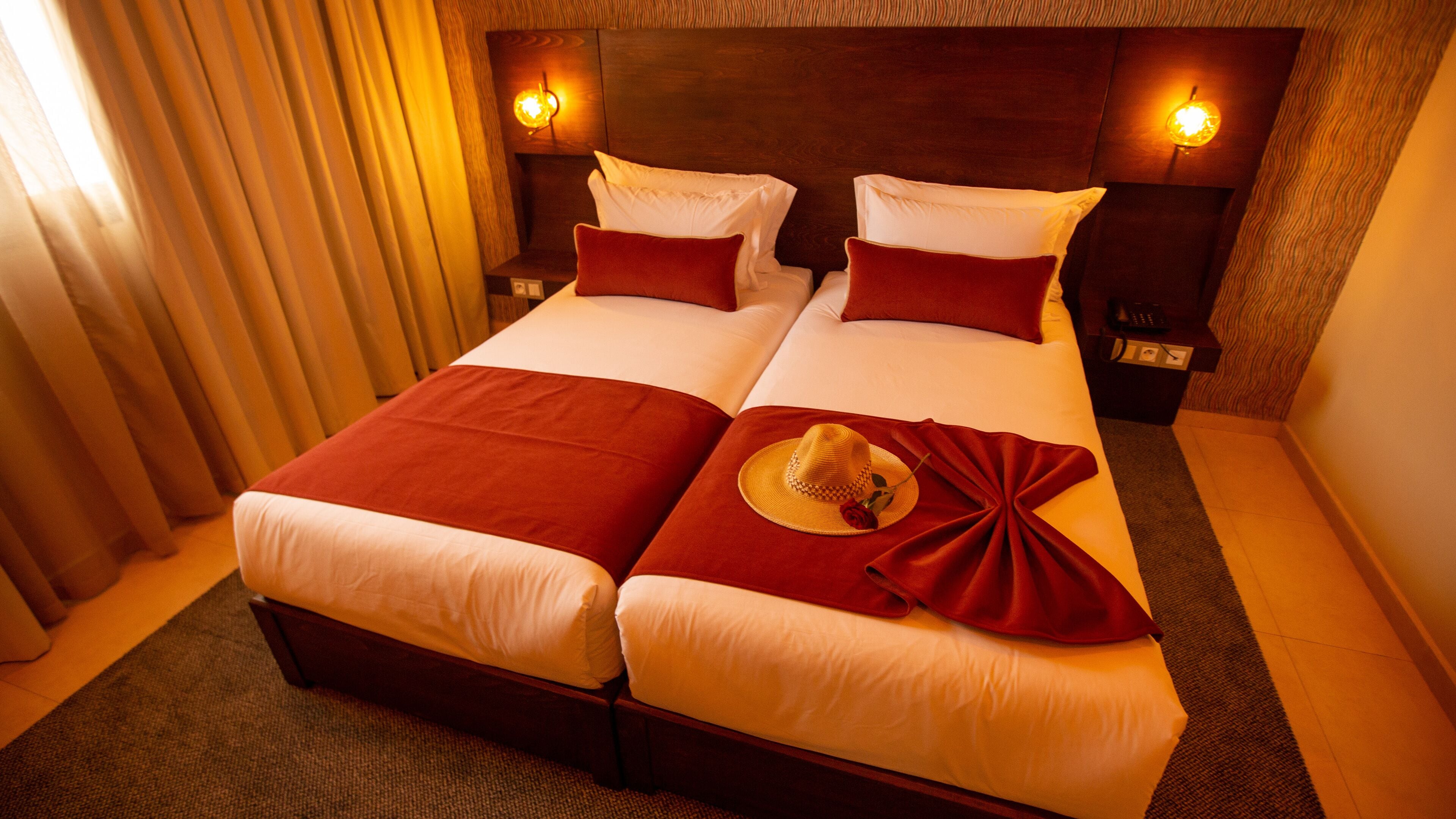 A double room with two beds, each with white linens and red accents. There are two bedside tables with lamps, a wooden headboard, and a large window with curtains.