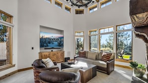 A spacious living room with a large TV, comfortable seating, and large windows offering a view of the outdoors.