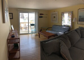 Uma sala de estar com sofá, mesa de centro, mesa de jantar com cadeiras, porta de vidro deslizante que dá para um pátio externo e um painel de parede estampado.