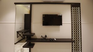 A modern living room with a mounted flat-screen TV, a built-in air conditioner, a small shelf with electronic devices, and a decorative partition. - Hotel Maple Inn (Ahmedabad)