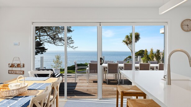 A modern kitchen with a view of the ocean, a dining area with outdoor seating, and a sign reading "BEACH HOUSE". - The Waters Edge by Ready Set Stay (San Remo)
