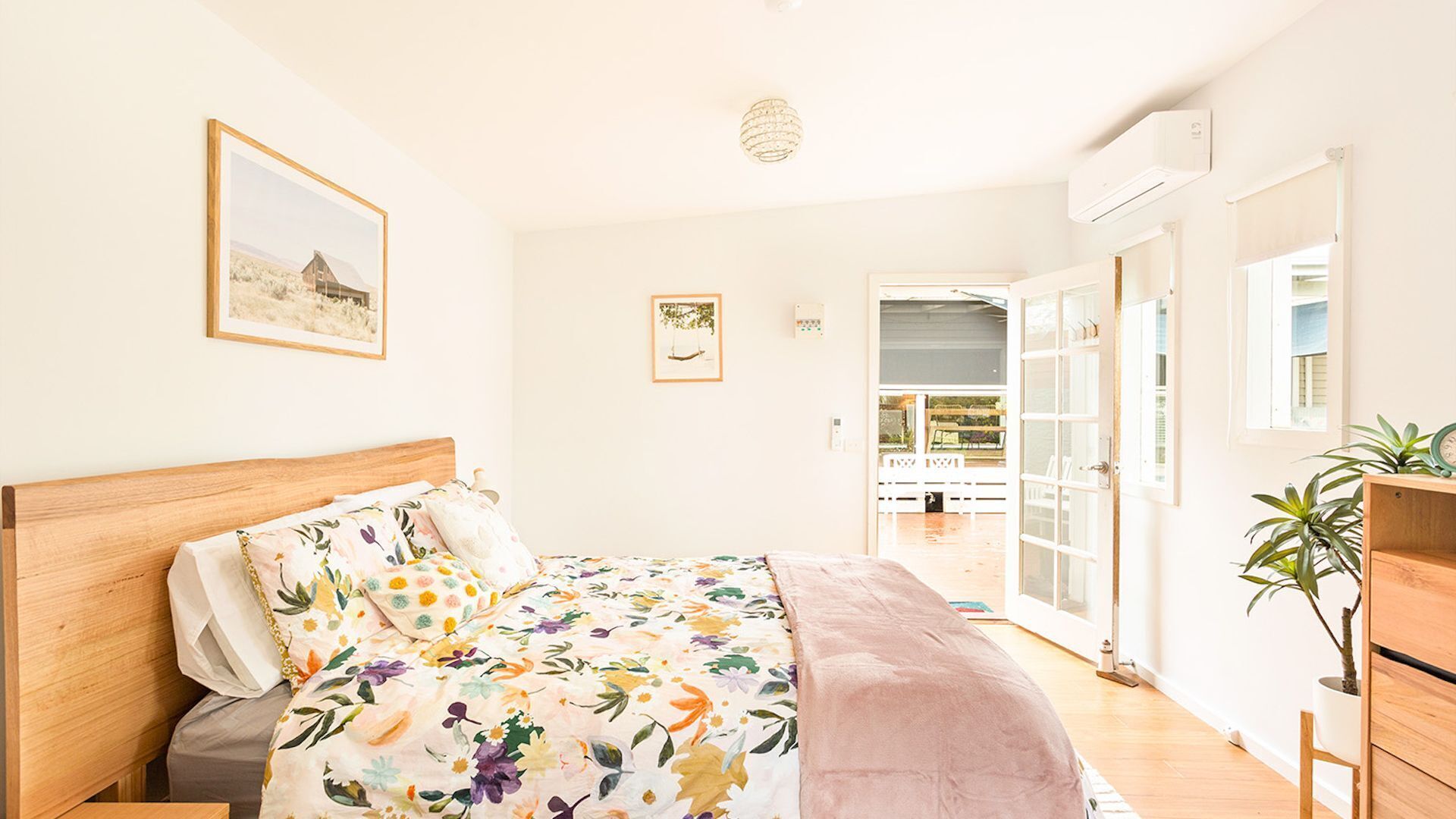 Quarto com cama de madeira, roupa de cama floral, vista para uma varanda externa e um quadro na parede.