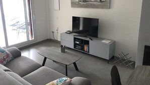 Una sala de estar moderna con un sofá gris, un televisor de pantalla plana sobre un mueble blanco, un cuadro con barcos en la pared, y un comedor con mesa y sillas.