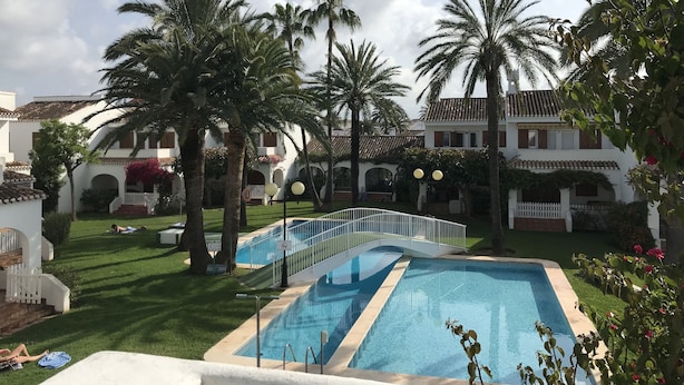 Zona de piscina con palmeras, cerca blanca y un edificio con techo de tejas al fondo.