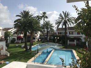 Zona de piscina con palmeras, cerca blanca y un edificio con techo de tejas al fondo.