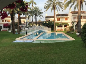 En swimmingpool med en hvid bro, omgivet af palmer og en velplejet græsplæne.