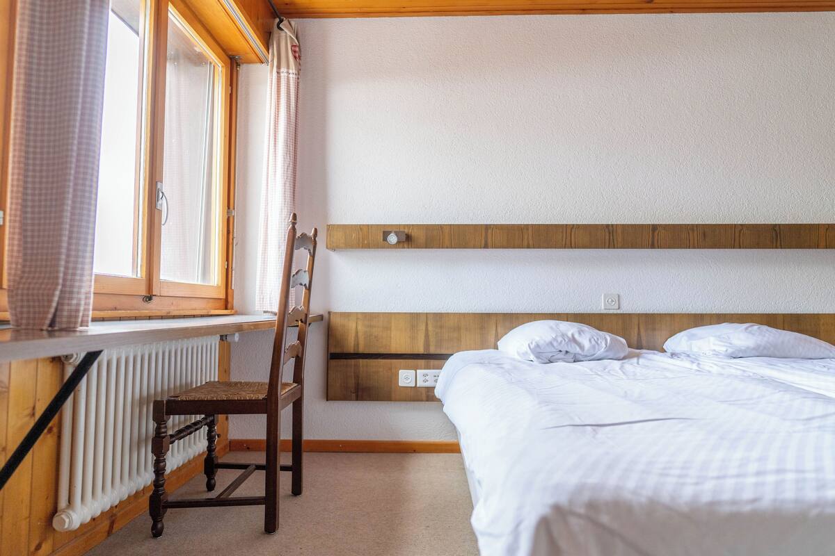 A bedroom with a wooden headboard, a single bed, a chair, a radiator, and a window with curtains.