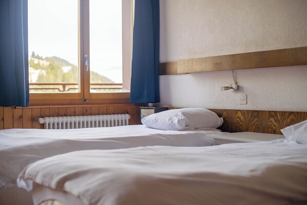 Quarto de hotel com cabeceira de madeira, cama branca, mesa de cabeceira e janela com cortinas.
