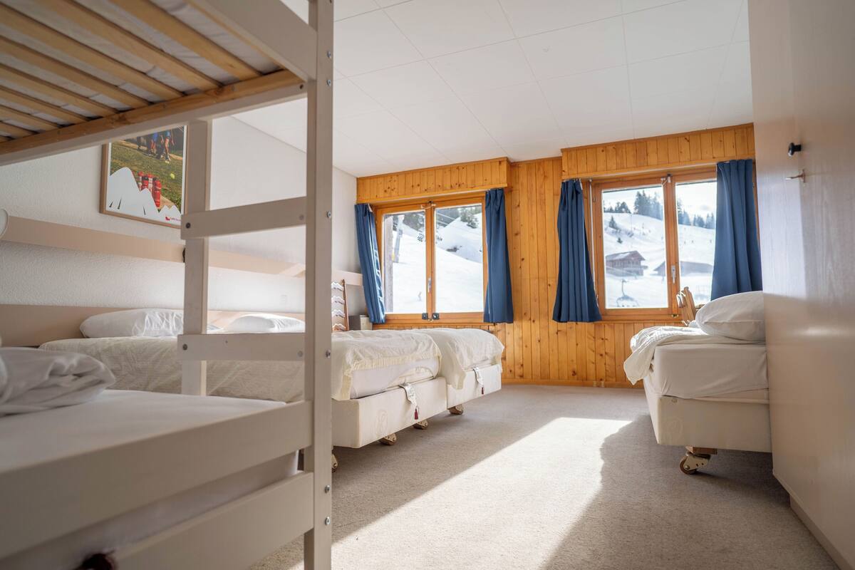 A cozy room with bunk beds, a wooden ceiling, large windows with curtains, and a view of a snowy landscape.