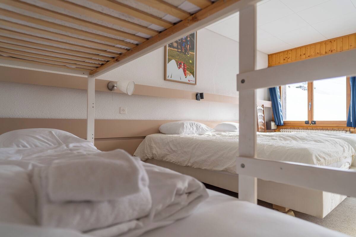 A bunk bed room with white bedding, a wooden ceiling, a framed picture on the wall, and a window with curtains.