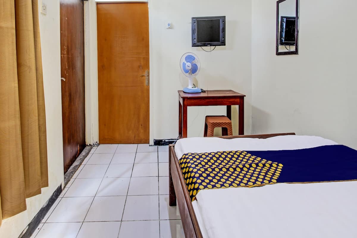 A hotel room with a bed, a wooden door, a small table with a fan, and a TV mounted on the wall.