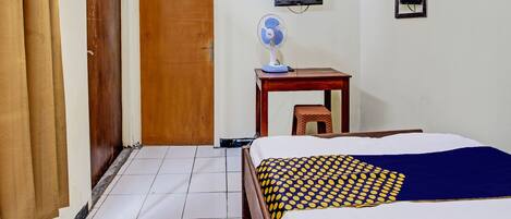 Habitación de hotel con cama, puerta de madera, una mesita con un ventilador y un televisor montado en la pared.