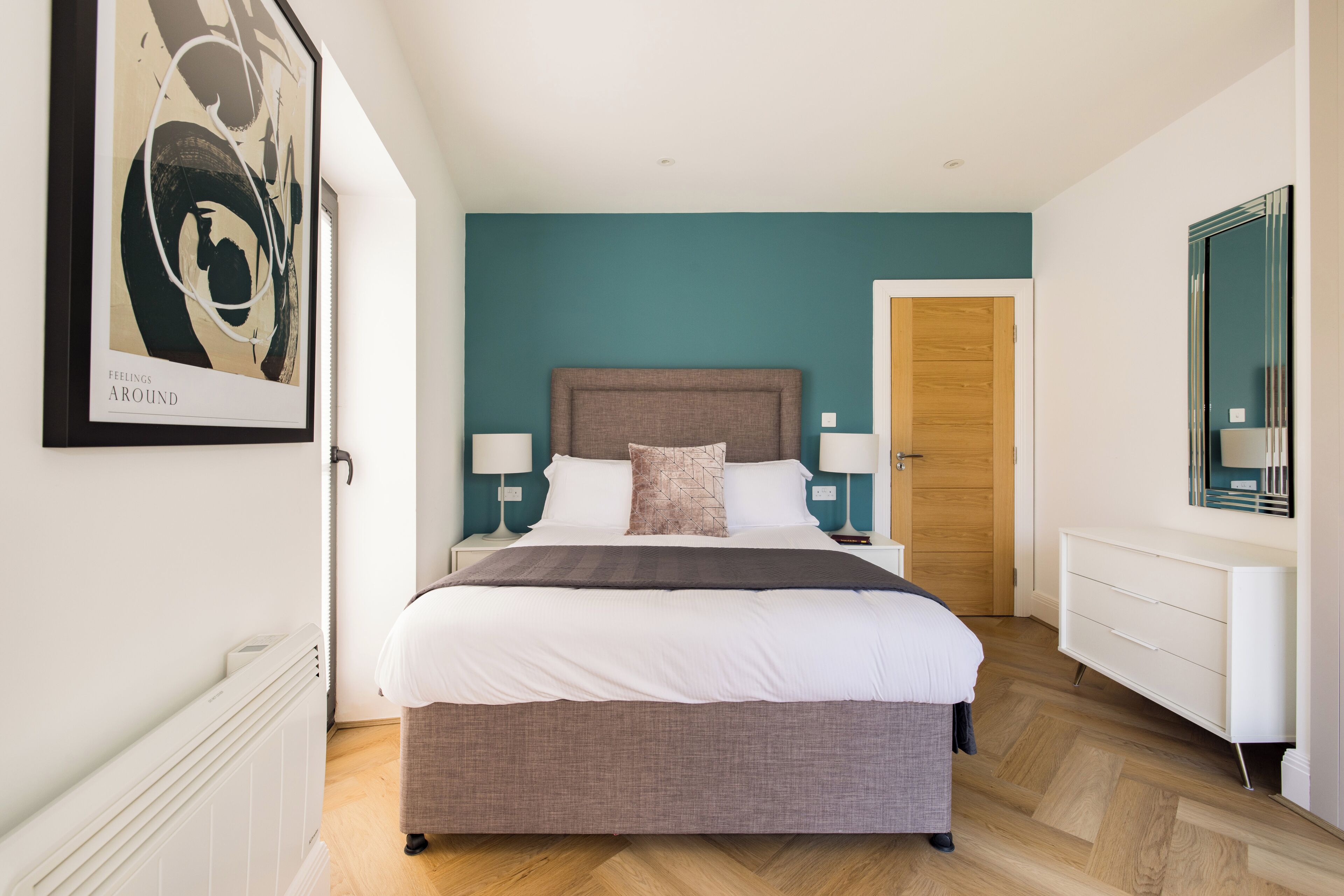 A modern bedroom with a large bed, two bedside tables with lamps, a wooden door, and a white dresser.