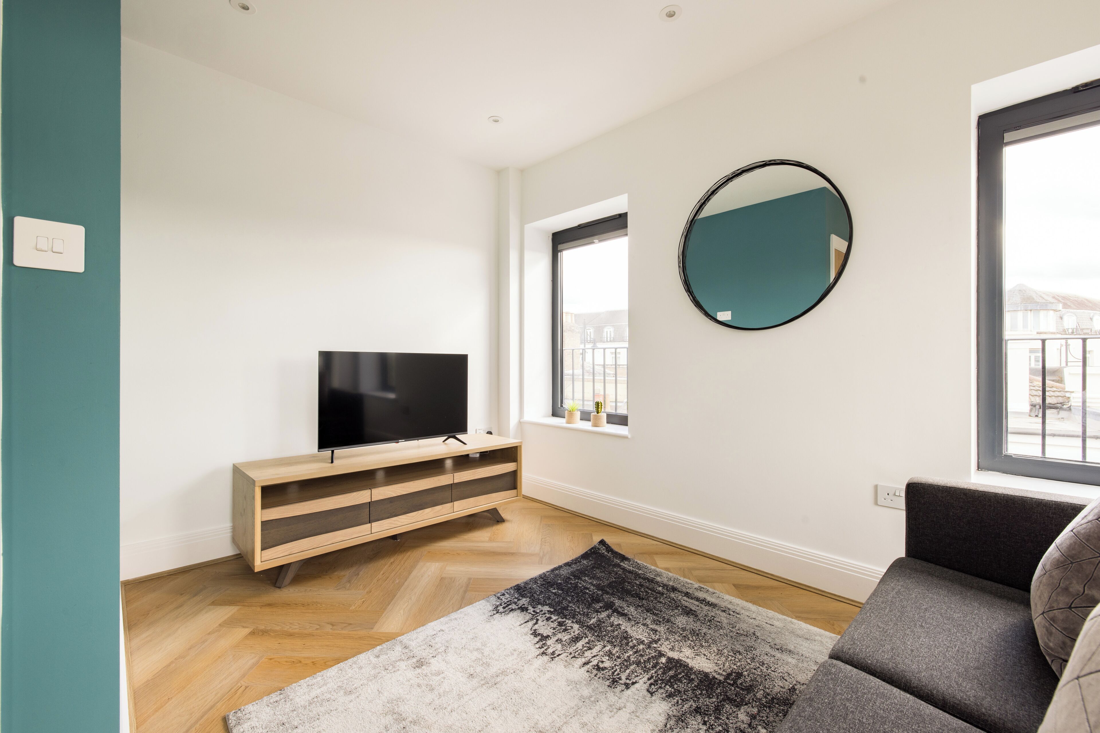 Una sala de estar moderna con un televisor de pantalla plana sobre un mueble de madera, un espejo grande en la pared y un sofá gris.